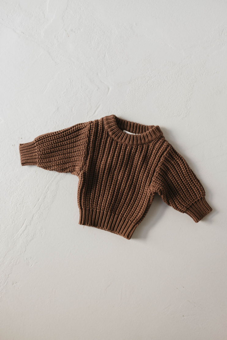 The Hot Cocoa Knit Sweater by forever french baby is a hand-knitted, chunky brown sweater for children with ribbed sleeves, cuffs, and hem, shown laid flat on a white textured surface.
