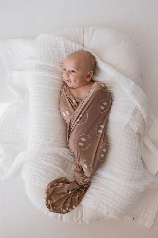 A baby is swaddled in a hypo-allergenic Bamboo Swaddle | Desert Clay ff Smile by forever french baby, lying on a white cushion. The baby is looking to the side with a calm expression. The white cushion and swaddle provide a soft and cozy environment.