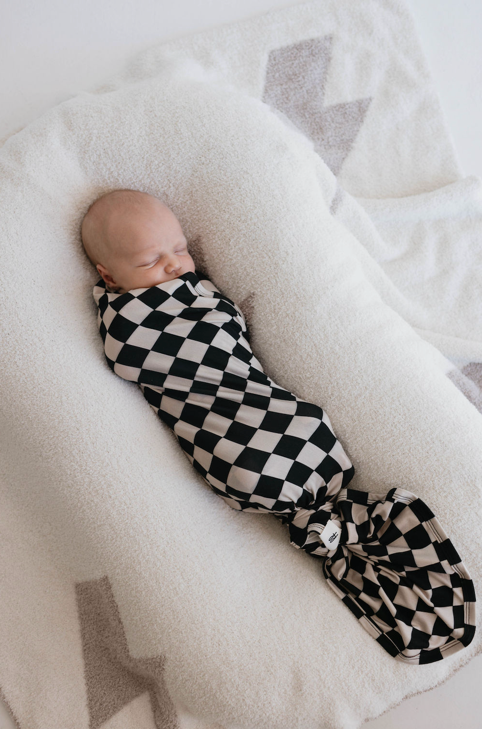 A baby is peacefully sleeping, wrapped snugly in a 'Black Checkerboard' hypo-allergenic bamboo swaddle from Forever French Baby. The baby is lying on a soft, white, cushioned surface with subtle gray geometric patterns visible on the fabric underneath.