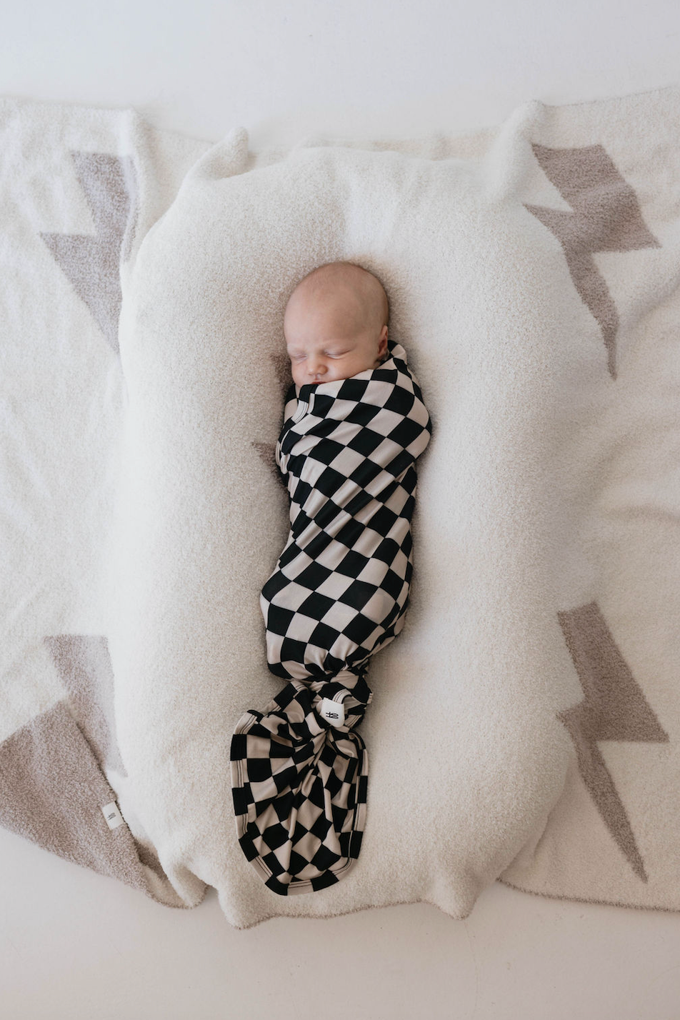 A baby wrapped in a Black Checkerboard Bamboo Swaddle from forever french baby is sleeping on a white plush blanket with light grey patterns on either side, lying peacefully in the center.