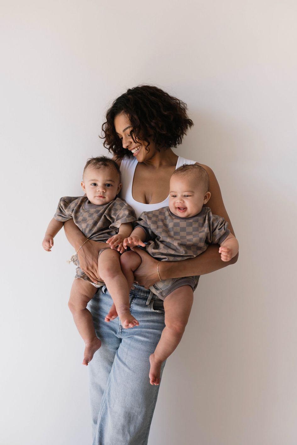 A woman with curly hair, wearing a white top and light blue jeans, smiles while holding two babies dressed in matching Forever French Baby Short Sleeve Rompers in Faded Brown Checkerboard. She looks at one of the babies as they pose against a plain white background.