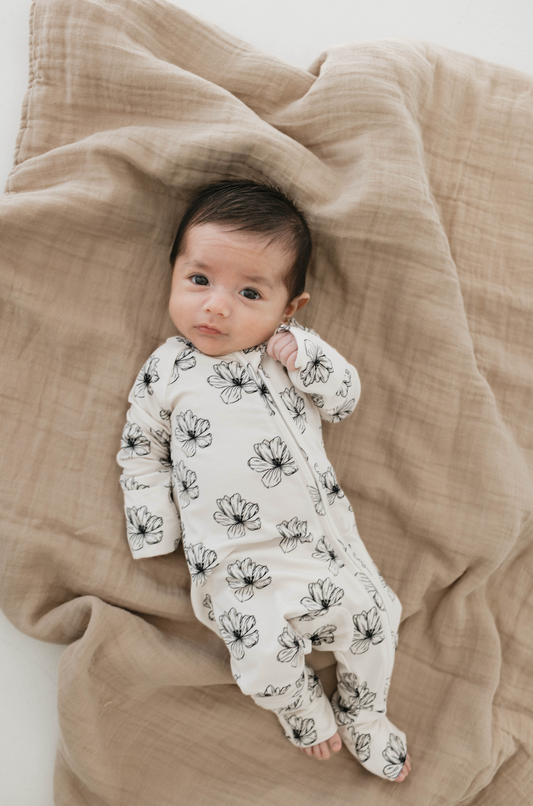 A baby in a Desert Bloom Bamboo Zip Pajamas from forever french baby lies on a soft, beige blanket. The baby, with dark hair and curious eyes, gazes upward, touching their cheek with one hand. The overall scene is cozy and serene.