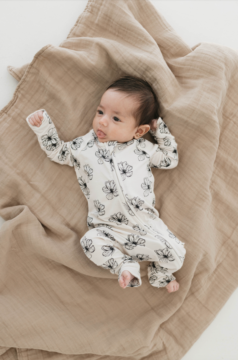 A baby wearing Forever French Baby's Bamboo Zip Pajamas in the Desert Bloom pattern lies on a soft beige blanket. The baby, with dark hair, looks up with one arm raised above their head and the other by their side. The setting is bright and minimalistic, perfectly highlighting the exquisite quality of Forever French Baby sleepwear.