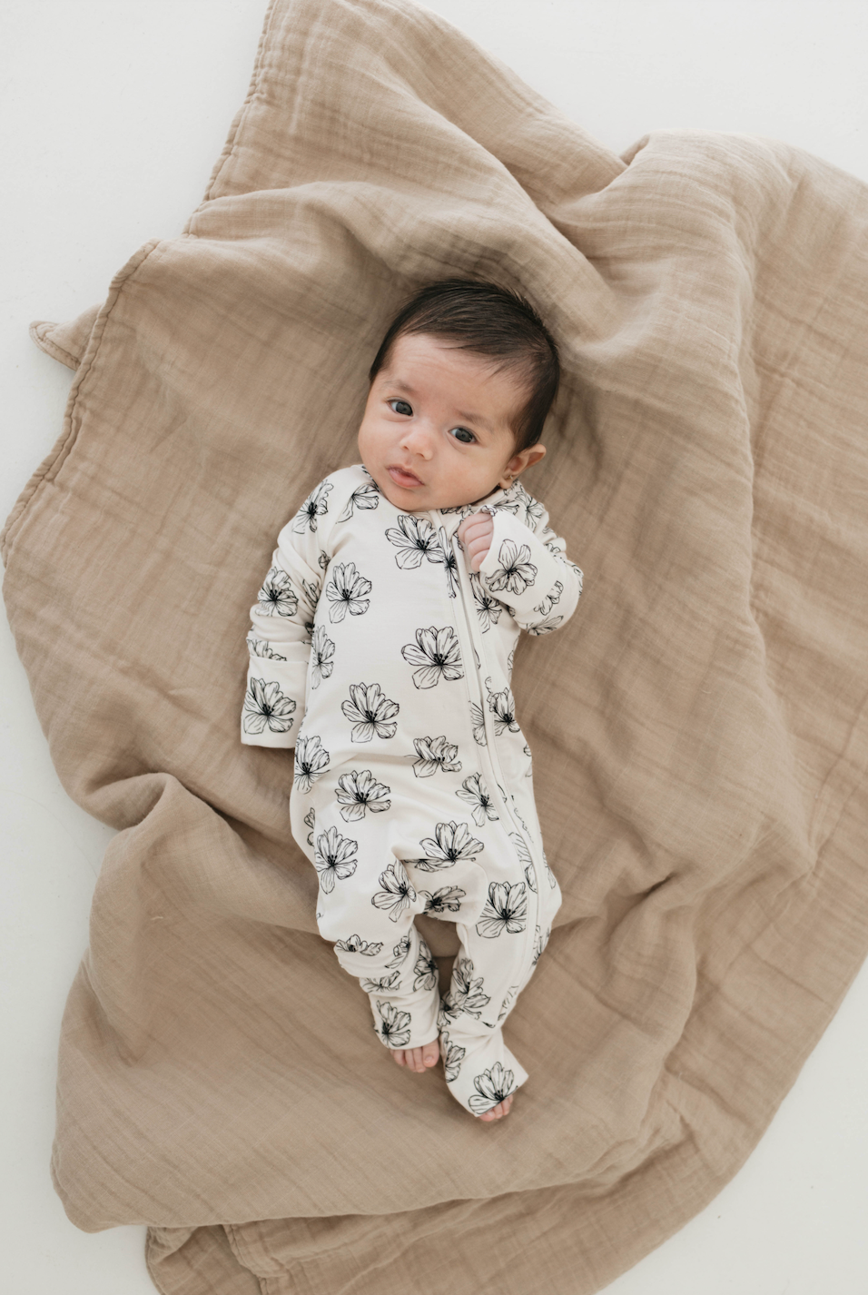 A baby dressed in Forever French Baby's Bamboo Zip Pajamas in the Desert Bloom pattern lies on a beige, textured blanket. The baby has short dark hair and is looking upwards with a neutral expression.