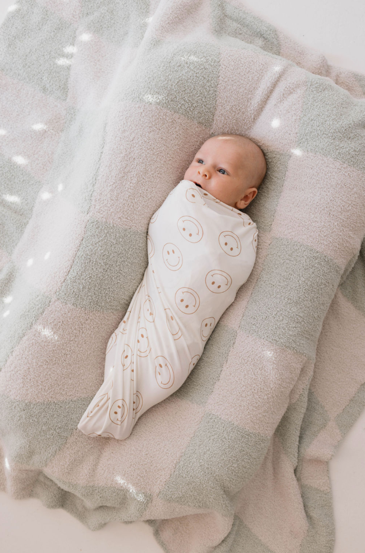 A baby wrapped in a forever French baby Bamboo Swaddle | Just Smile, adorned with smiley face designs, lies peacefully on top of a soft, checkered blanket featuring light pink and gray squares. The hypo-allergenic baby clothing ensures the infant appears calm and content while gazing upwards.