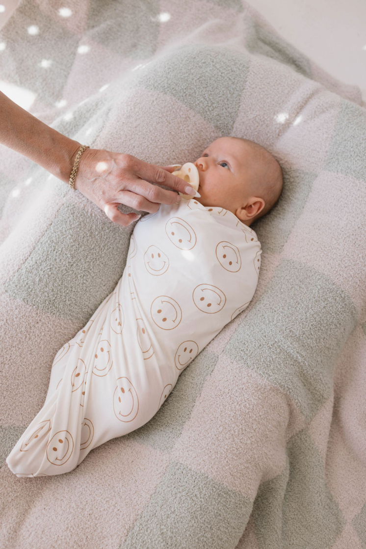 A baby cozily wrapped in a forever French baby Bamboo Swaddle | Just Smile with smiley face patterns lies on a soft, checkered blanket. An adult hand gently holds a pacifier to the baby's mouth as light filters through, creating a dappled effect on the scene, ensuring year-round comfort for the little one.