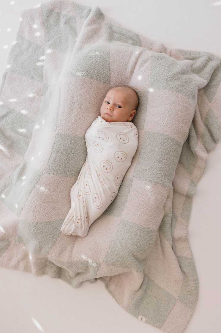 A baby swaddled in a "Just Smile" ivory bamboo swaddle by forever French baby, featuring smiley face patterns, lies atop a soft, checkered blanket in pastel pink and mint green. As the baby looks up, patches of sunlight create a dappled effect on the blanket, providing year-round comfort.