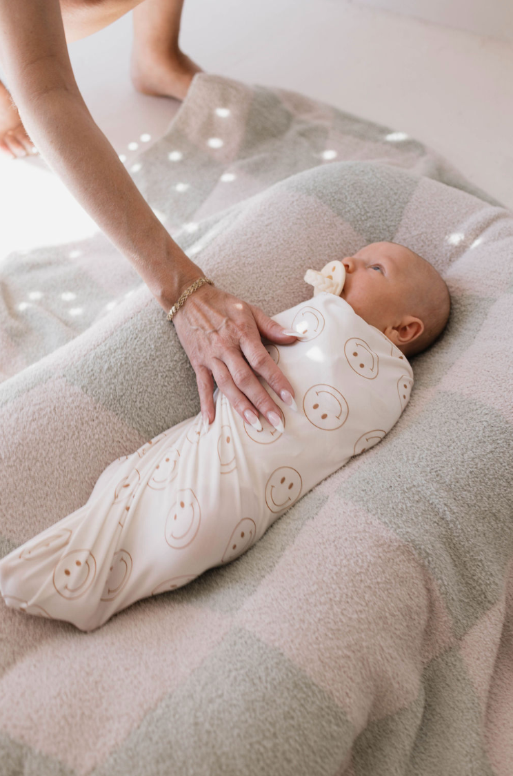A baby wrapped in a Forever French Baby "Just Smile" Bamboo Swaddle lies on a soft pink and gray checkered blanket. The baby has a pacifier in their mouth while an adult's hand gently rests on the baby's chest. Light filters through a nearby window, casting a soft glow that ensures year-round comfort.