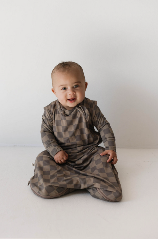 A baby wearing a long-sleeved, brown and beige checkered outfit is sitting on a white surface against a plain background, smiling and looking directly at the camera. This precious moment highlights the importance of baby's sleep needs, making the Bamboo Sleep Sack in Faded Brown Checkerboard from forever french baby an ideal choice for comfort.