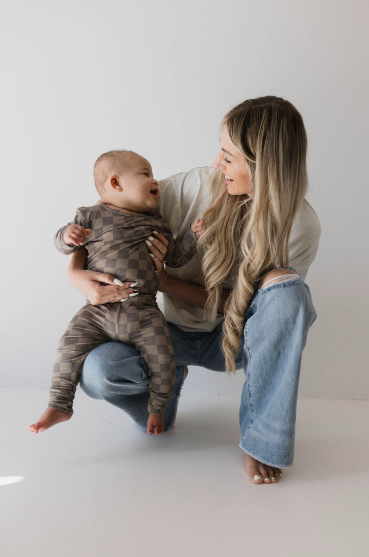 A woman with long blonde hair, dressed in a white t-shirt made of breathable bamboo fabric and blue jeans, kneels and laughs with a baby. The baby, wearing the Sleepy Time Set in Faded Brown Checkerboard by Forever French, looks up at the woman with a smile. The background is plain white.
