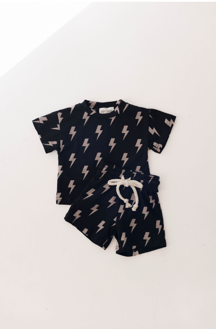 The "Short Set | Black Lightning" by forever french baby is a children's clothing set designed for ages 0-9 years. This outfit includes a dark blue short-sleeved shirt and matching shorts, both adorned with a pattern of black and white lightning bolts. The shorts feature a white drawstring at the waist for easy adjustment, all set against a plain white background.