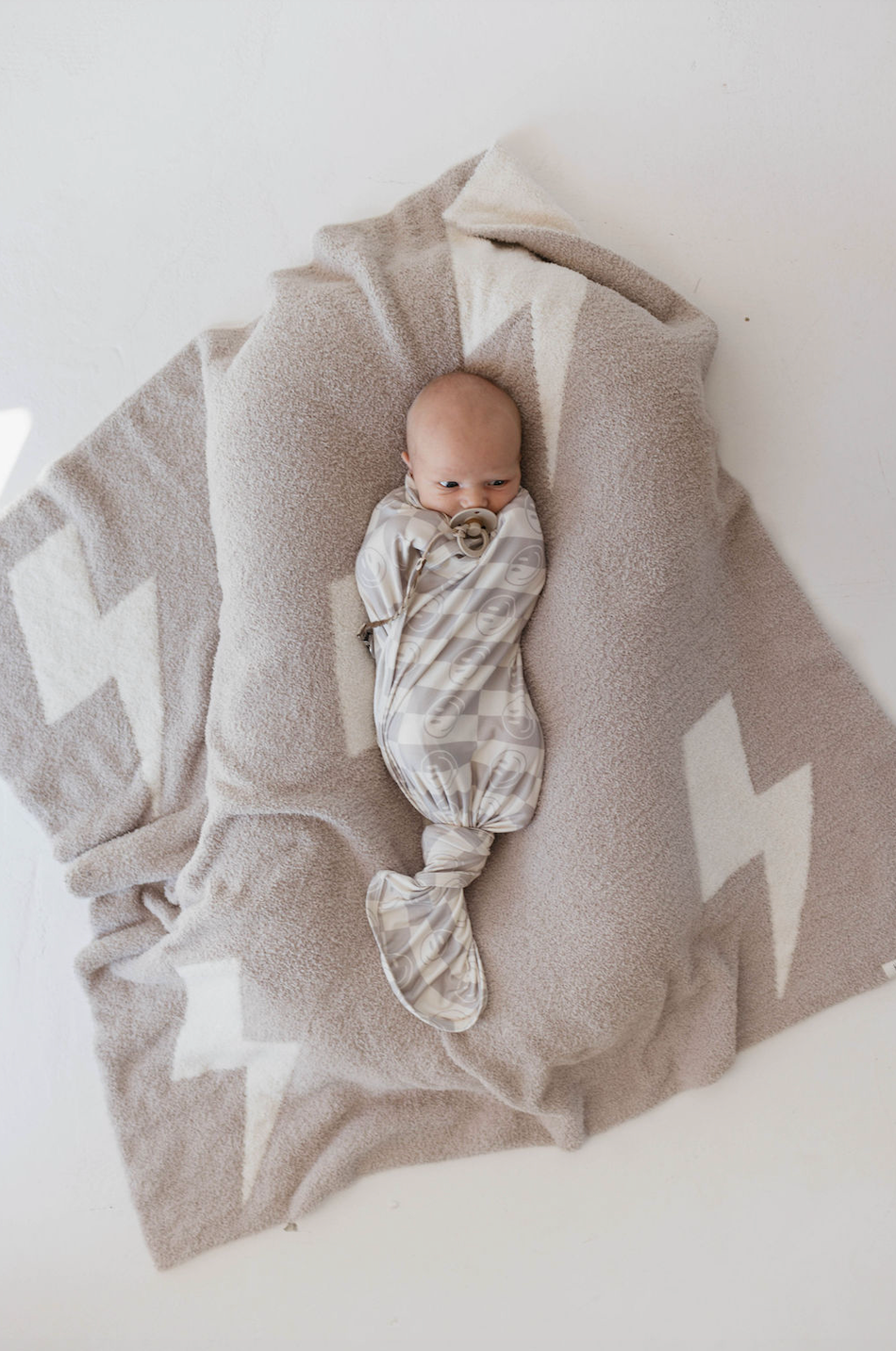A baby is swaddled in a Plush Blanket | Monsoon by forever french baby, featuring light gray and white geometric patterns and made from Oeko-tex certified materials. The baby is laying on a soft beige blanket adorned with white lightning bolt designs, resting on a white surface as they look directly at the camera.
