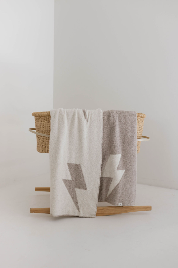 A simple, modern wicker bassinet features two "Plush Blankets | Monsoon" from forever french baby draped over its side. One blanket is white with a beige lightning bolt pattern, and the other is beige with a white lightning bolt pattern. The bassinet stands on a wooden rocking frame against a minimalist white background.