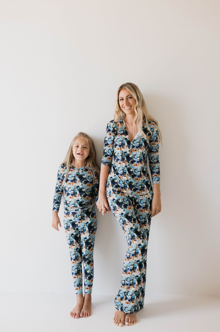 A woman and a young girl stand barefoot against a white wall, both wearing matching forever french baby Women's Bamboo Pajamas in the Charli Print. They are holding hands and smiling warmly, appearing relaxed and happy.