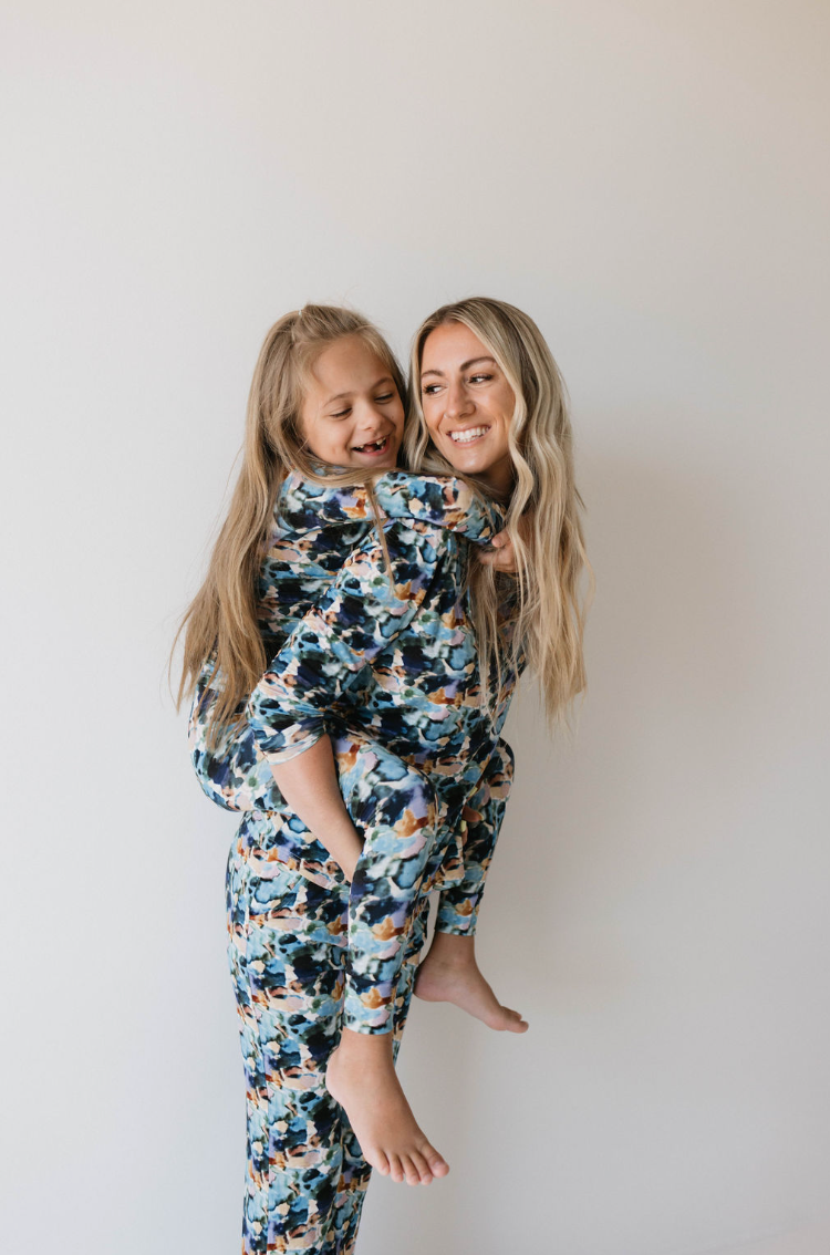 A joyful woman with long blonde hair is smiling as she carries a young girl on her back. Both are wearing matching Charli Print bamboo pajamas from forever french baby, featuring colorful floral designs. The backdrop is a plain white, highlighting their happiness and the girl's happy gaze towards the woman, promoting autism awareness through their loving bond.