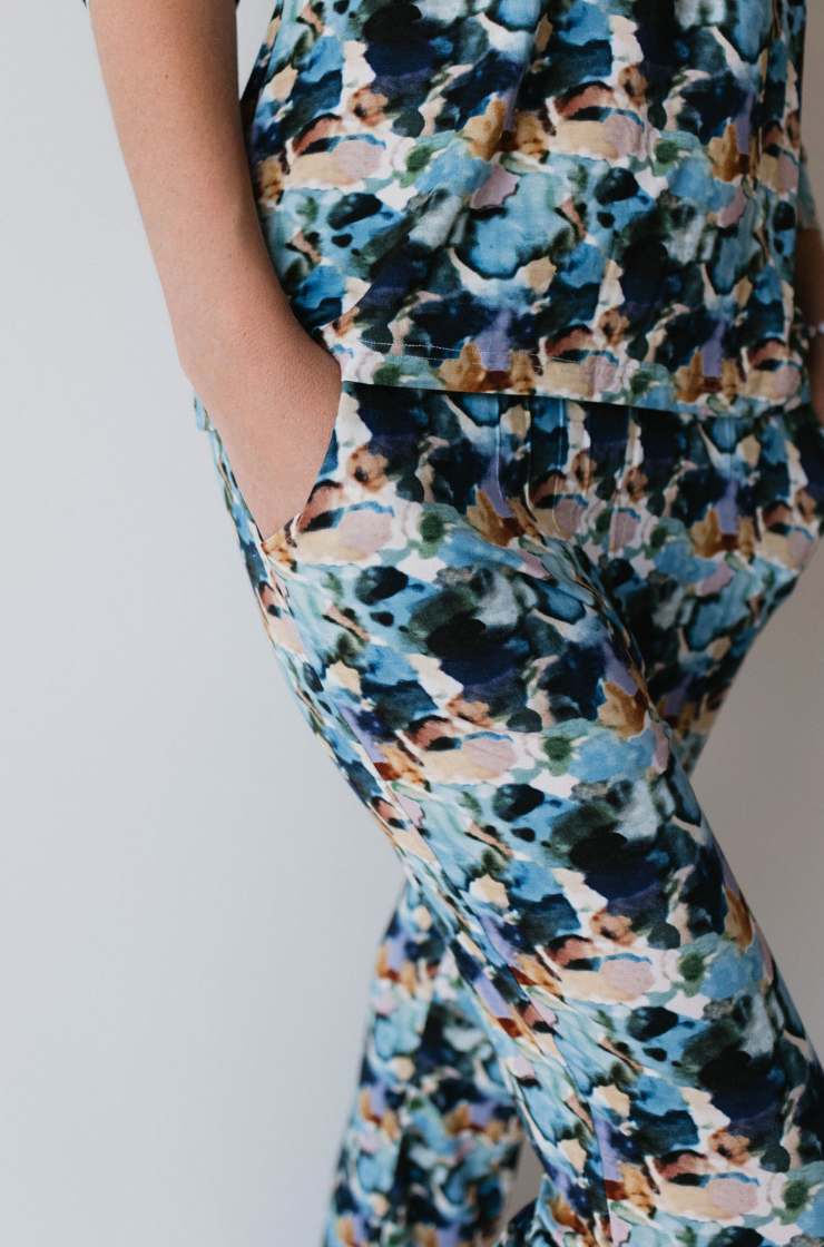 Partial view of a person wearing forever french baby's Women's Bamboo Pajamas in the Charli Print, featuring an abstract and colorful pattern in shades of blue, green, beige, and black. The person has one hand in their pocket and is standing against a plain white background. Only the midsection and legs are visible.