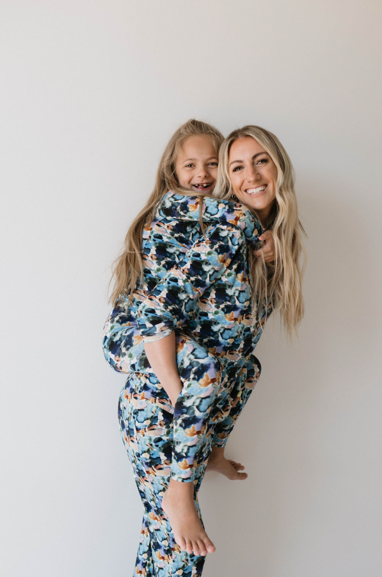 A woman with long blonde hair, dressed in a colorful floral-patterned outfit from forever french baby, smiles while piggybacking a young girl wearing matching Women's Bamboo Pajamas | Charli Print. They stand against a plain, light-colored background.