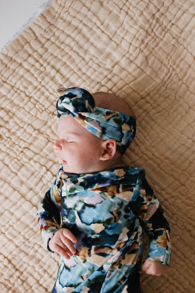 A baby sleeps peacefully on a quilted light brown blanket, dressed in a colorful Charli Print Bamboo Knotted Gown from forever french baby with a matching headband. The image captures a serene and cozy atmosphere, reminiscent of the gentle comfort found in bamboo baby gowns.
