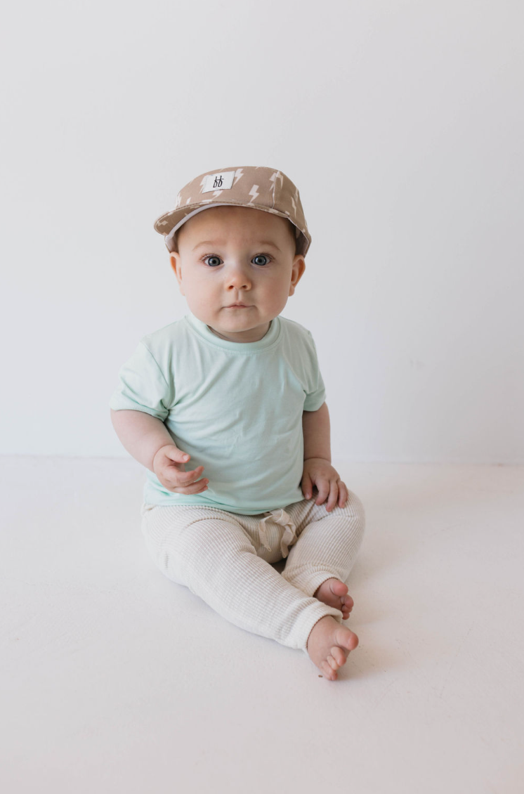 A baby wearing a mint Basic Bamboo Tee from forever french baby, beige pants, and a camo-patterned cap sits on a white floor against a white background. The baby is looking directly at the camera with a neutral expression, and one hand is raised slightly.