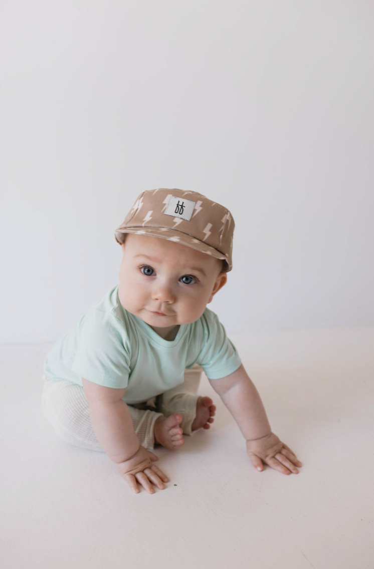 A baby wearing a mint-colored Basic Bamboo Tee from forever french baby, white pants, and a brown hat with a lightning bolt pattern sits on a white floor against a white background, looking up with wide eyes.