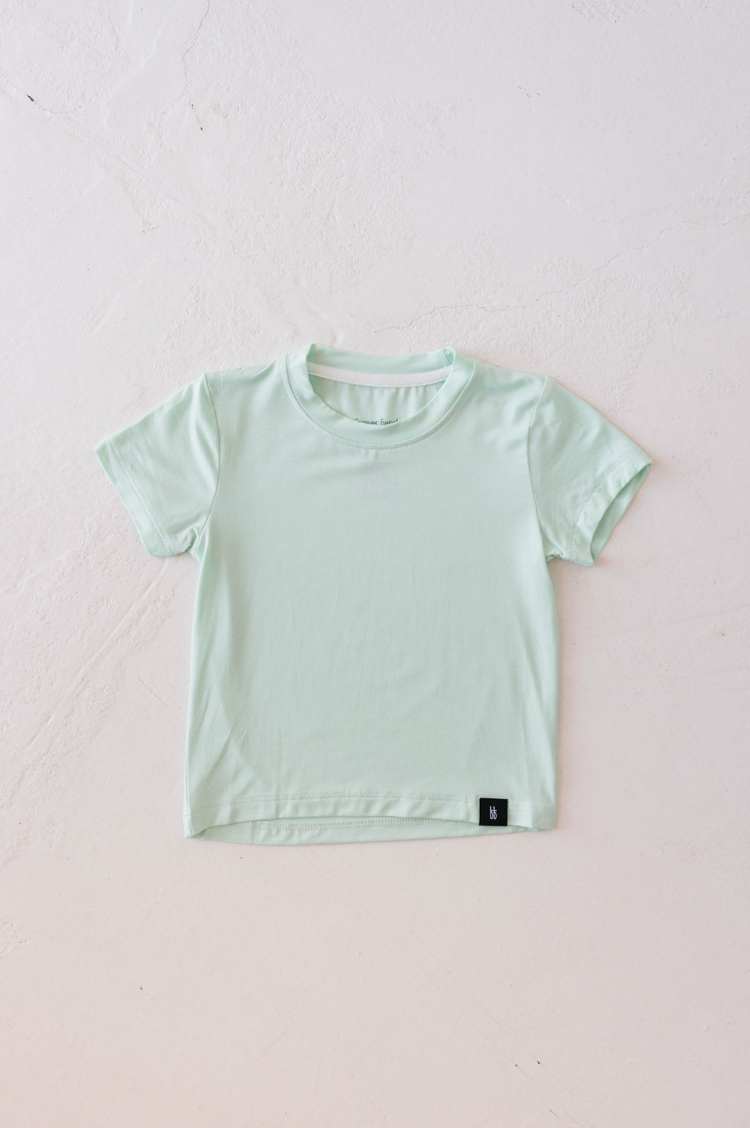 A Basic Bamboo Tee | Mint from forever french baby is neatly laid out on a white textured surface. Made from soft bamboo fabric, the shirt has a simple design with a small black tag on the bottom hem.