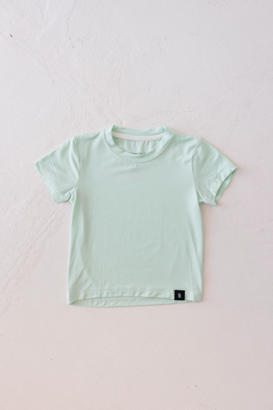 A Basic Bamboo Tee | Mint from forever french baby is neatly laid out on a white textured surface. Made from soft bamboo fabric, the shirt has a simple design with a small black tag on the bottom hem.