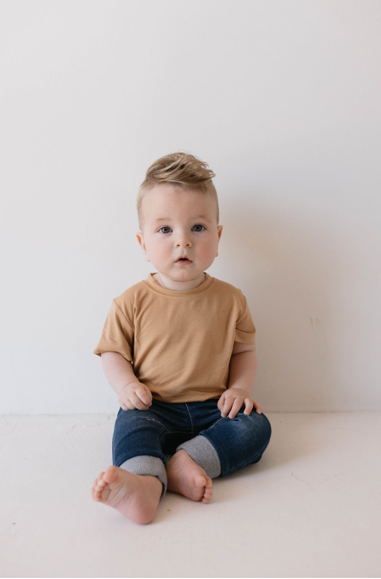 A young child with short, light hair sits against a white background. The child is wearing the Basic Bamboo Tee | Desert Sunset from Forever French Baby paired with dark blue jeans featuring gray cuffs. Made from soft bamboo fabric, the shirt ensures the baby is comfortable. The child looks towards the camera with a neutral expression.