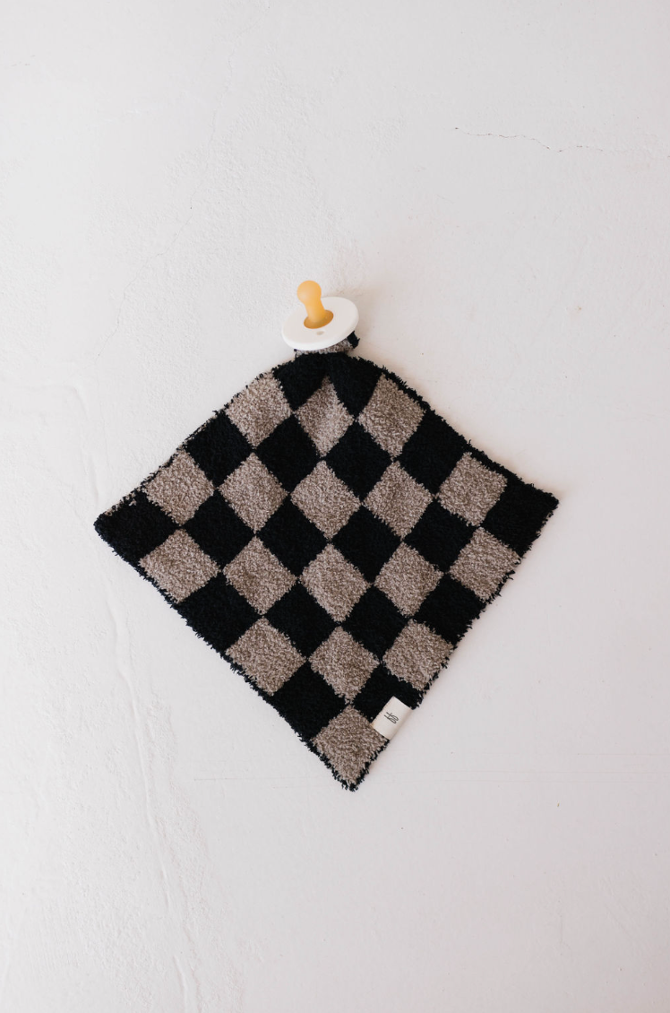 The "Lovey | Checkmate Checkerboard" by forever french baby, a luxurious microfiber feather yarn security blanket in a black and white checkerboard pattern, elegantly graces a light-colored surface. It features an attached wooden ring and pacifier at one corner—an exquisite creation for your little one.