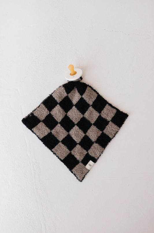 The "Lovey | Checkmate Checkerboard" by forever french baby, a luxurious microfiber feather yarn security blanket in a black and white checkerboard pattern, elegantly graces a light-colored surface. It features an attached wooden ring and pacifier at one corner—an exquisite creation for your little one.
