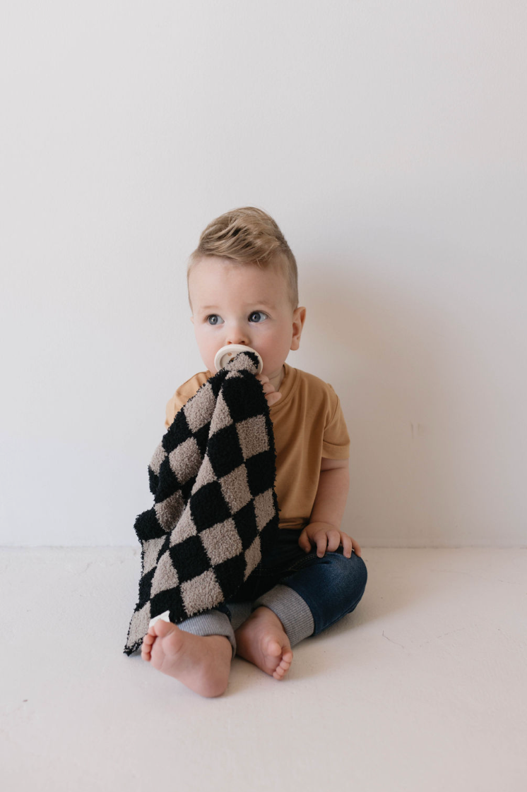 A small child with short blond hair sits against a white wall, wearing a tan shirt and blue jeans. The child is holding the Lovey | Checkmate Checkerboard blanket from forever french baby and has a pacifier in their mouth, looking slightly to the side.