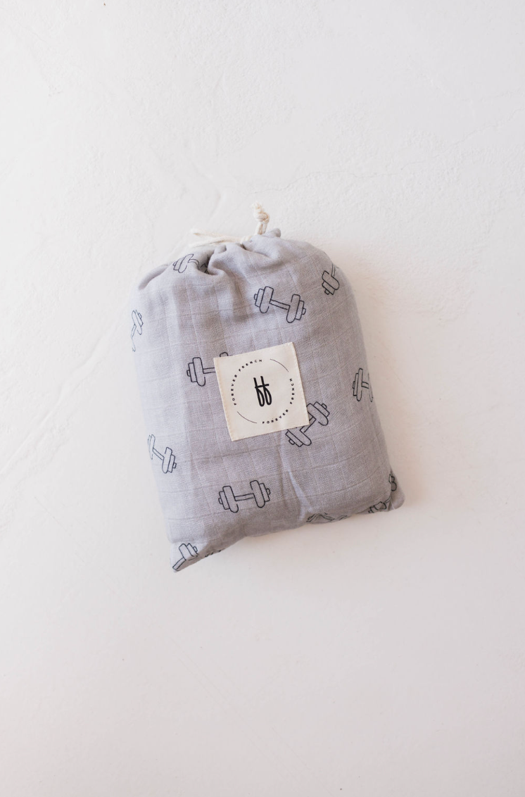 A muslin crib sheet named "Arm Day" by Forever French Baby, featuring a light gray fabric with a black dumbbell design and adorned with a white square patch displaying the brand's logo of two lowercase "b" letters in black, is laid on a white surface.