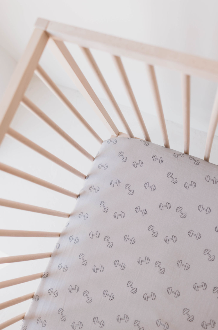 Top view of a wooden baby crib with the "Muslin Crib Sheet | Arm Day" by forever french baby, featuring pale grey bedding adorned with a small, evenly spaced dumbbell design. The crib has simple, widely spaced wooden bars, creating a clean and minimalistic setting ideal for modern nursery decor.