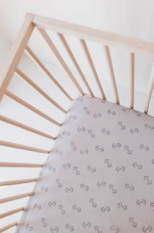 Top view of a wooden baby crib with the "Muslin Crib Sheet | Arm Day" by forever french baby, featuring pale grey bedding adorned with a small, evenly spaced dumbbell design. The crib has simple, widely spaced wooden bars, creating a clean and minimalistic setting ideal for modern nursery decor.