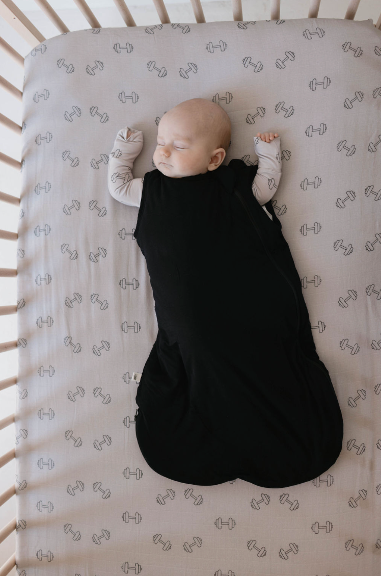 A baby sleeps peacefully in a crib, wearing a black sleep sack over light-colored clothes. The crib bedding, part of the charming nursery decor from forever french baby, features the Muslin Crib Sheet | Arm Day with a pattern of small, gray dumbbells. The baby's arms are raised, and the crib rails are shown on each side.