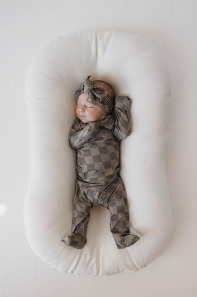 A sleeping newborn dressed in the Forever French Sleepy Time Set in Faded Brown Checkerboard, made from breathable bamboo clothing with a matching headband, lies on a white cushioned baby sleeper. The baby has its arms raised and appears peaceful and content.