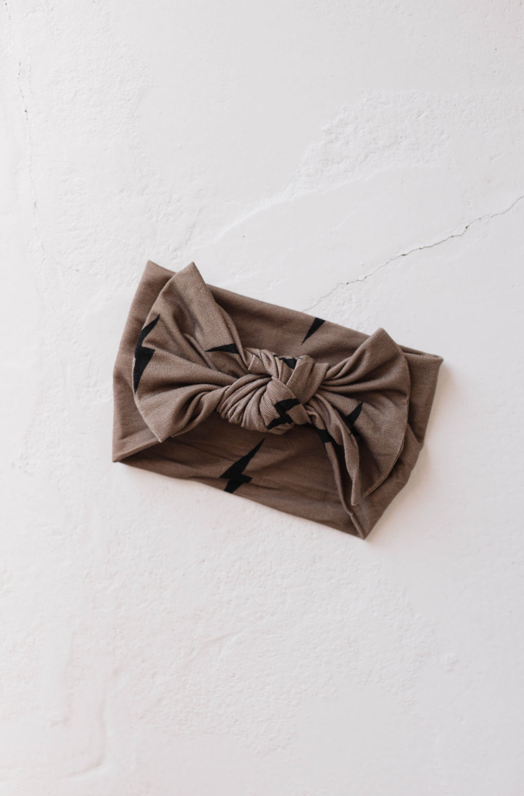 The Bamboo Head Wrap in Brown & Black Bolt from forever french baby, featuring a large bow and black zigzag pattern, is elegantly placed on a textured off-white surface. This chic accessory adds a touch of elegance and charm to any outfit.