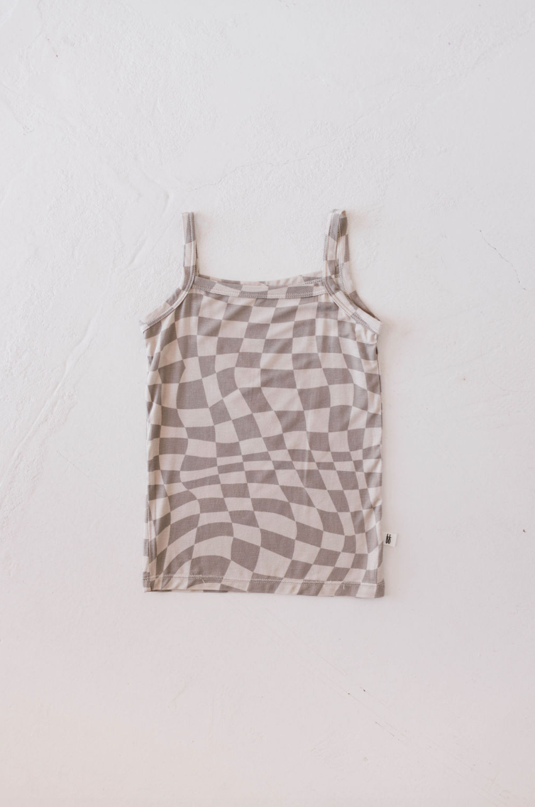 The Basic Bamboo Tank | Wavy Checker from forever french baby, featuring a beige and light brown checkered pattern with a wavy, distorted design, is laid flat on a white textured surface. Crafted from soft bamboo fabric, the tank top includes thin shoulder straps and a small tag on the seam for ensuring all-day comfort for your little one.