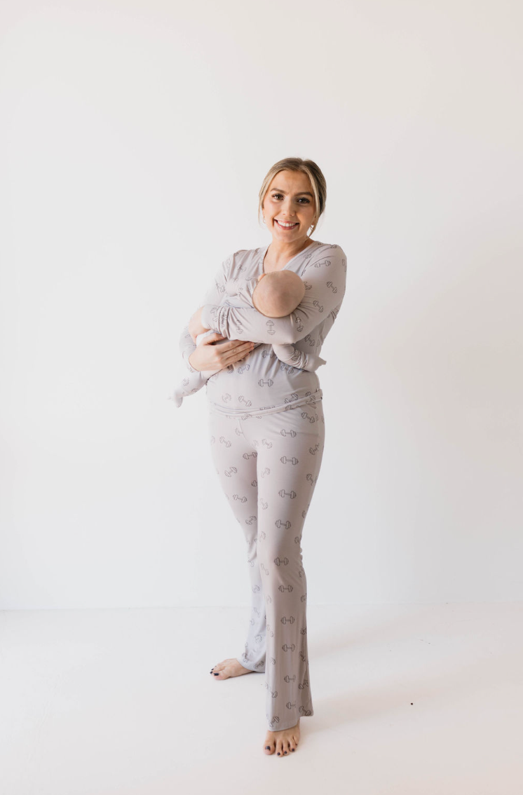 A woman stands barefoot against a white background, smiling at the camera. She is wearing matching gray Women's Bamboo Pajamas | Arm Day from forever french baby with a subtle pattern and is cradling a baby wrapped in a similar gray outfit, which blends harmoniously with her attire.