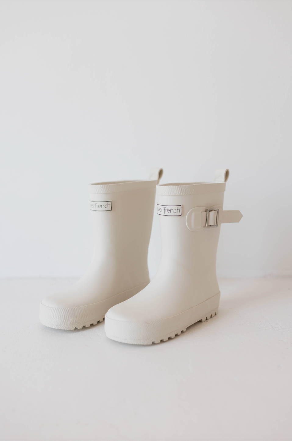A pair of ff Rain Boots in cream from forever french baby. These off-white rubber children's rain boots feature a buckled strap on the outer sides and have a sturdy sole with lug patterns for traction. The boots are placed on a light surface against a plain white background.