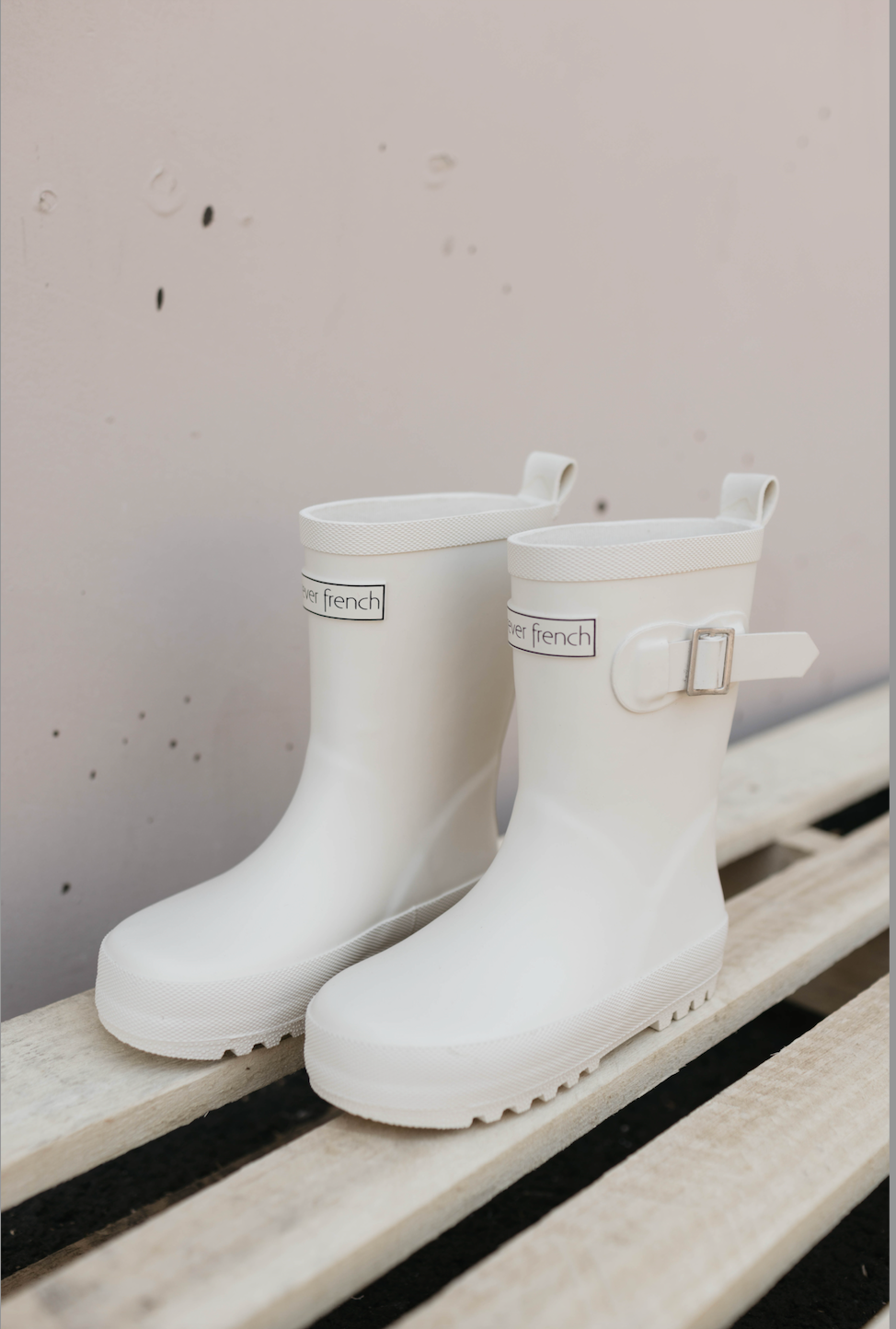 A pair of the ff Rain Boots in Cream from forever french baby, with silver buckles on the sides, placed on a light-colored wooden pallet against a light pink wall. These all-weather kids' footwear feature a loop at the back for easy wearing and have "very french" text near the top edges.