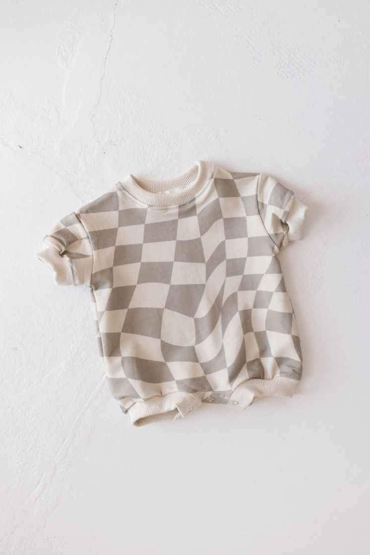 The Forever French Baby Short Sleeve Romper, featuring a beige and cream wavy checkerboard design, is displayed flat on a light-colored surface. Made from organic cotton, this romper includes ribbed cuffs and snap buttons at the bottom for easy changing.