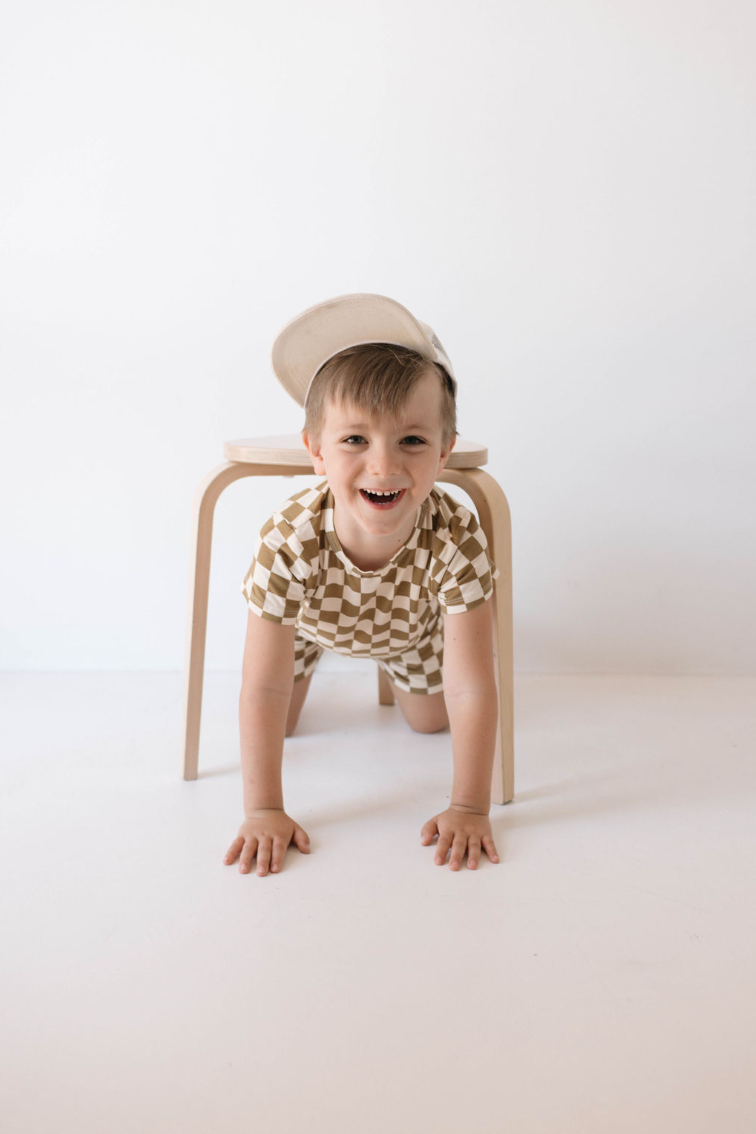 A young child with short brown hair, dressed in the breathable Bamboo Short Set in Olive Checkerboard from forever french baby and a white cap, smiles while crawling under a wooden chair in a white room.