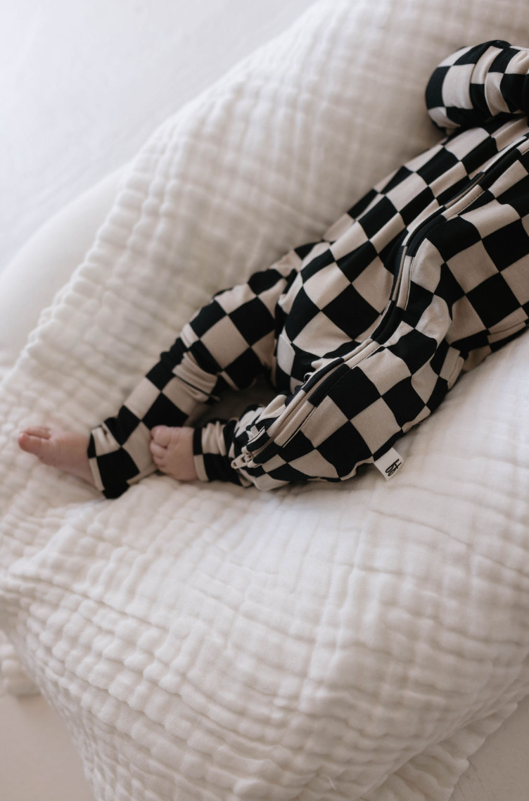 A baby wearing Forever French Baby's Bamboo Zip Pajamas in Black Checkerboard with rollover cuffs is lying on a soft, white textured blanket. The baby's feet are visible, and the positioning suggests a peaceful and relaxed state.