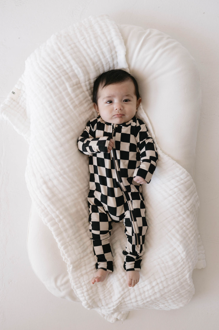 A baby lies on a soft white cushion, wearing Bamboo Zip Pajamas in Black Checkerboard from forever french baby. The baby has dark hair and gazes upward with a calm expression. The cushion is wrapped in a textured white blanket, adding to the cozy ambiance of the scene.