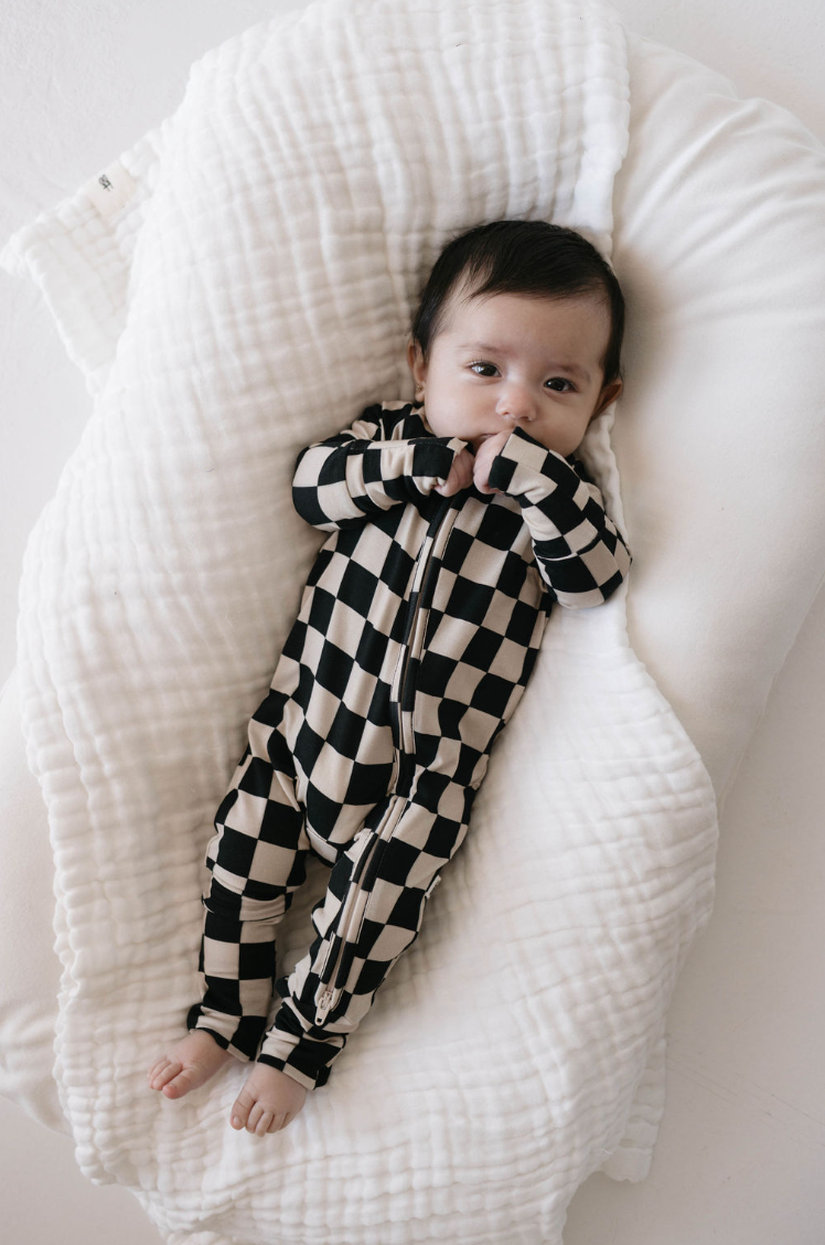 A baby with dark hair lies on a soft, white blanket. The baby is wearing Forever French Baby's Bamboo Zip Pajamas in a black checkerboard pattern, with their left hand partially in their mouth. The baby gazes up, appearing calm and comfortable.