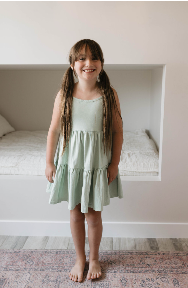 A young girl with long brown hair and bangs stands smiling in a room. She is wearing the forever french baby Children’s Dress in Vintage Washed Spearmint, featuring a tiered skirt. Her hair is tied in two low ponytails. Behind her is a bed recessed into the wall with white bedding.