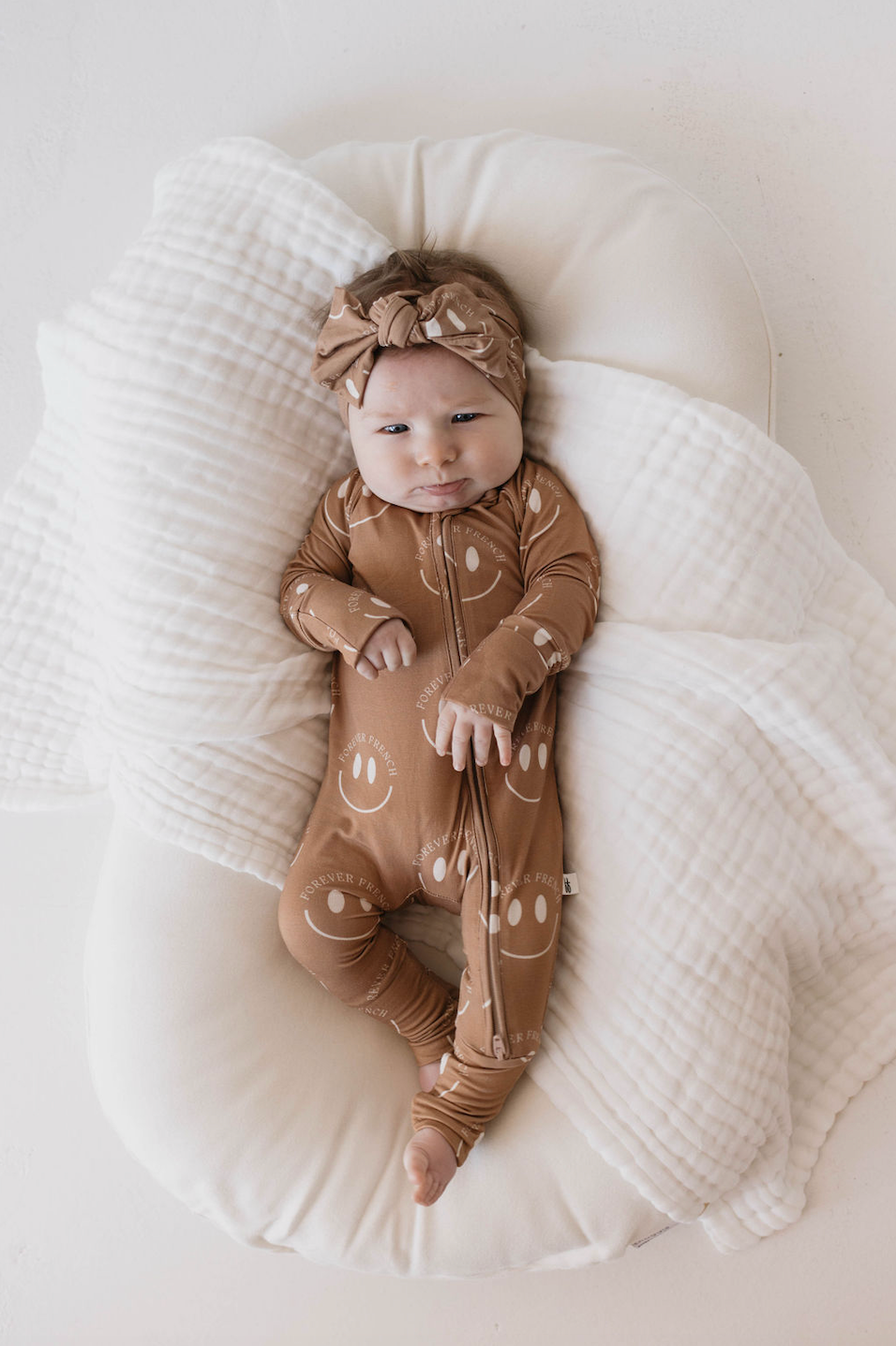 A baby wearing a "Bamboo Head Wrap | ff Desert Clay Smile" from Forever French Baby lies on a soft, white cushioned surface, warmly wrapped in a white textured blanket made from bamboo material.
