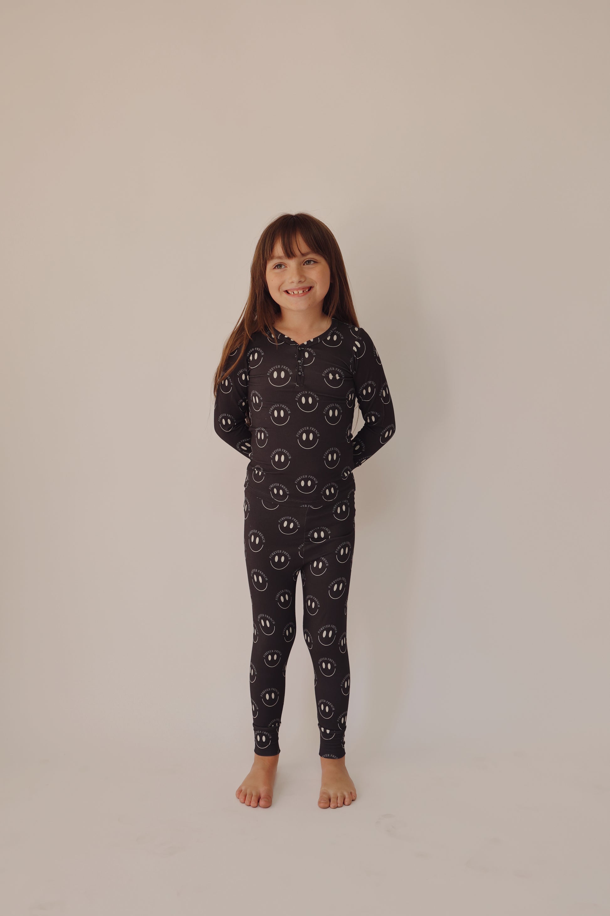 A young child with long brown hair stands barefoot, smiling and wearing the Bamboo Two Piece Pajamas in Charcoal & White ff Smile by forever french baby. The child stands against a plain off-white background.