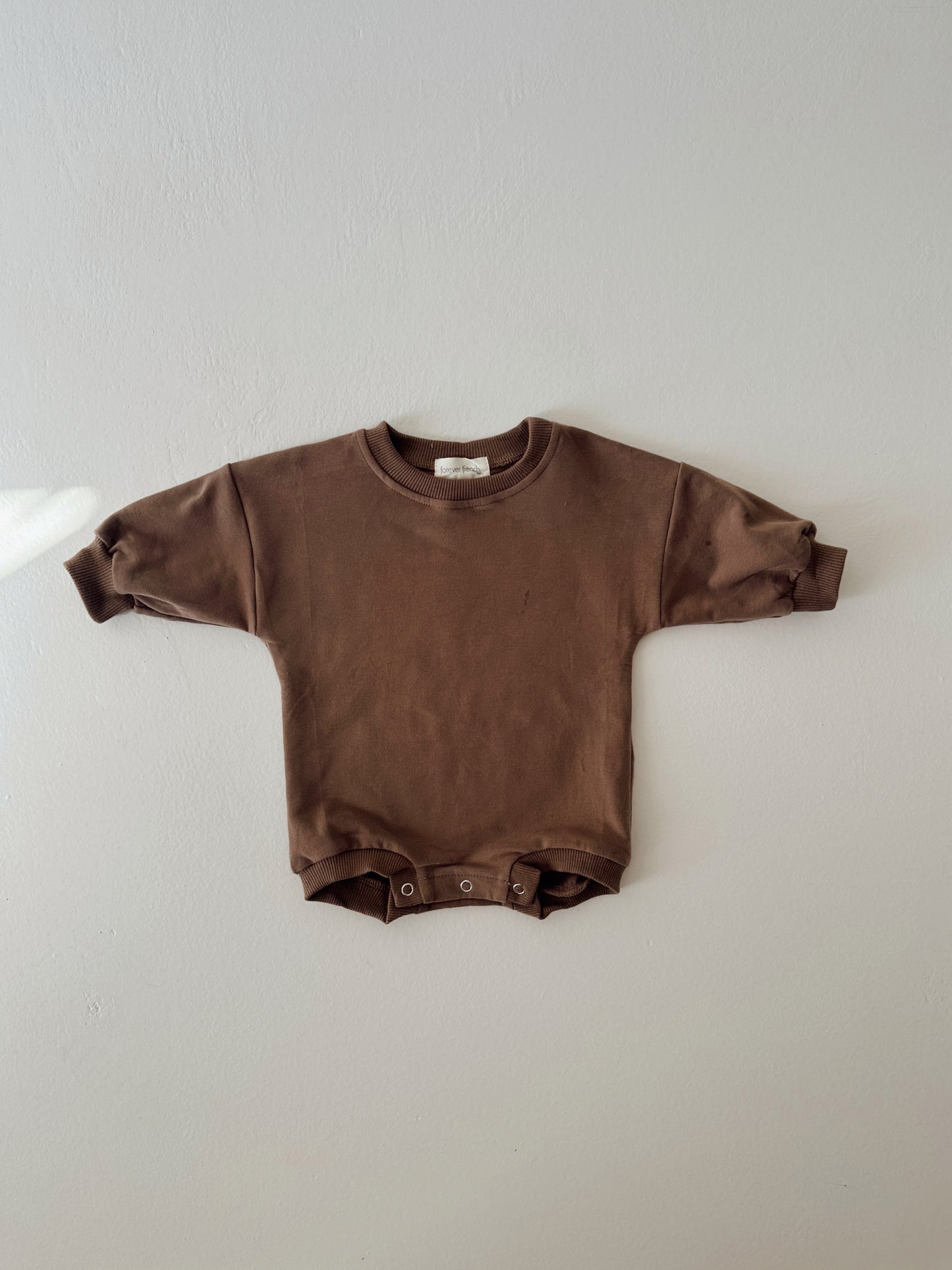 The forever french baby Sweatshirt Romper | Hot Cocoa, an organic cotton brown romper with long sleeves and snap buttons at the crotch, is displayed flat against a light gray wall.
