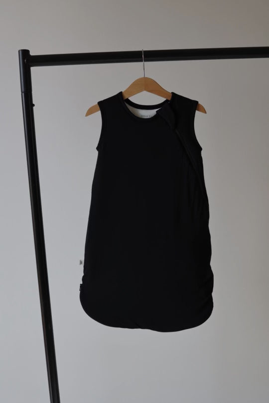A Bamboo Sleep Sack in Midnight Black by forever french baby with a round neckline hangs on a wooden hanger against a minimalist backdrop. Made from breathable bamboo fabric, the sleeveless sleep sack features a simple design and is displayed on a black garment rack.